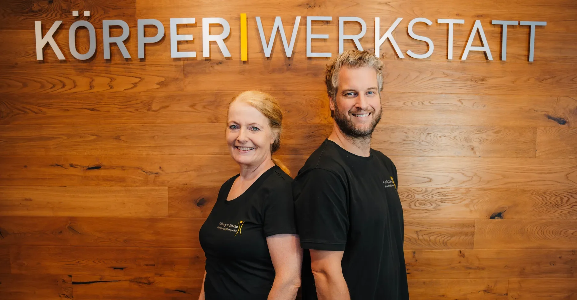 Zwei Mitglieder des Körperwerkstatt-Teams stehen Rücken an Rücken vor der hölzernen Wand mit dem Schriftzug Körperwerkstatt.